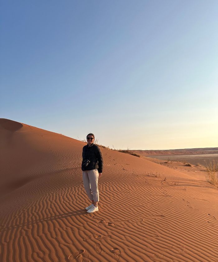 A person stands on a desert sand dune under a clear blue sky. The sun casts soft shadows, highlighting the ripples in the sand.