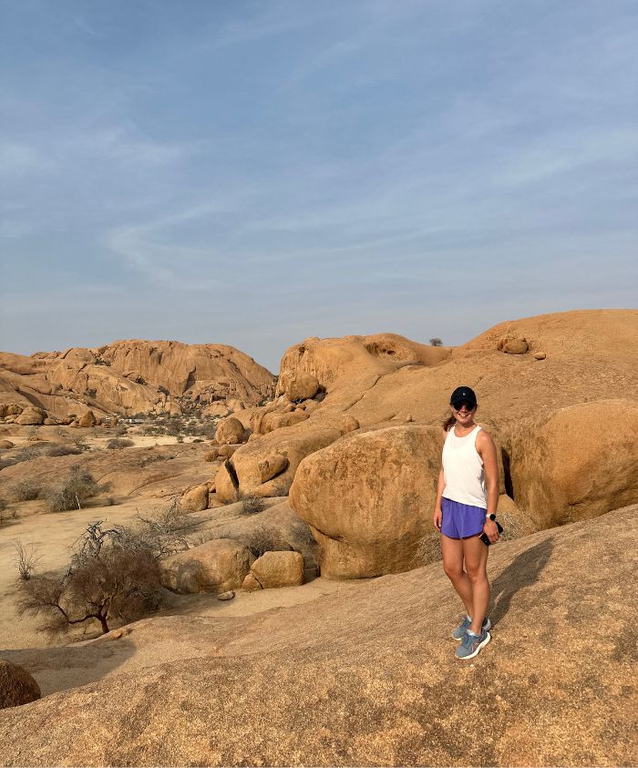 A person wearing a white tank top, purple shorts, and sneakers stands on a rocky landscape with large boulders and a dry terrain. The sky is clear with a hint of clouds.