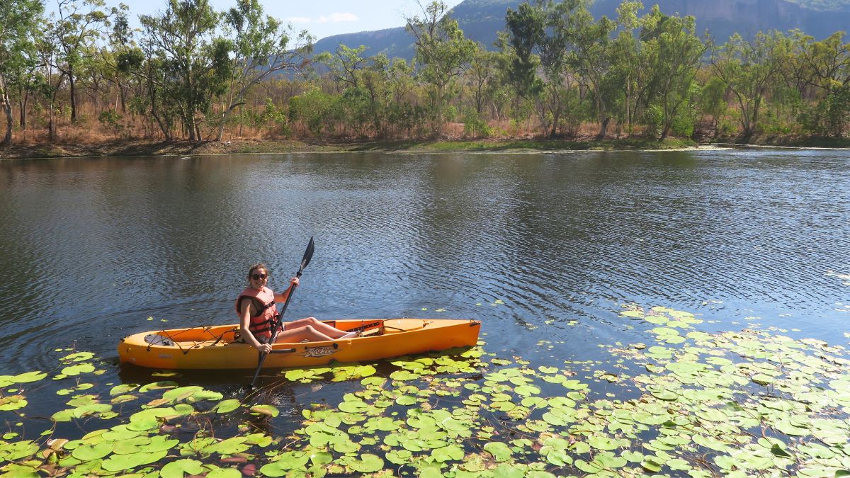A person kayaking in a bright yellow kayak, surrounded by lily pads on a calm river. Lush green trees line the bank, with distant hills in the background.