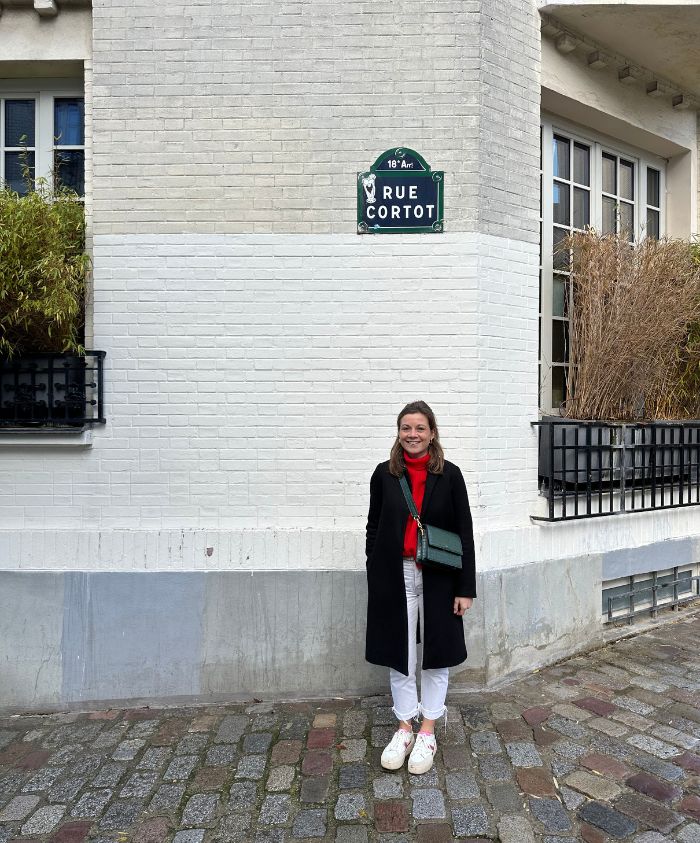 A woman stands in front of a white brick wall with a street sign reading "Rue Cortot." She wears a black coat, red scarf, and white trousers, with her hands in her pockets.