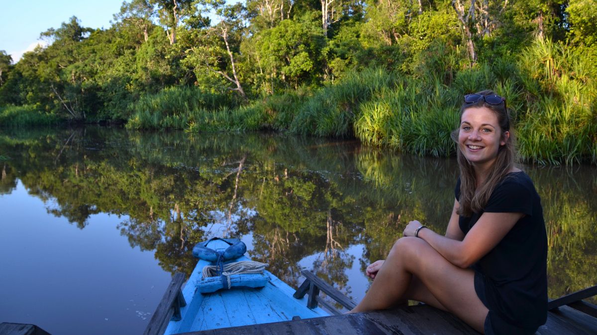 A young woman with long brown hair sits on the edge of a blue wooden boat, smiling at the camera. The serene river reflects the lush greenery surrounding it, creating a peaceful, natural scene.