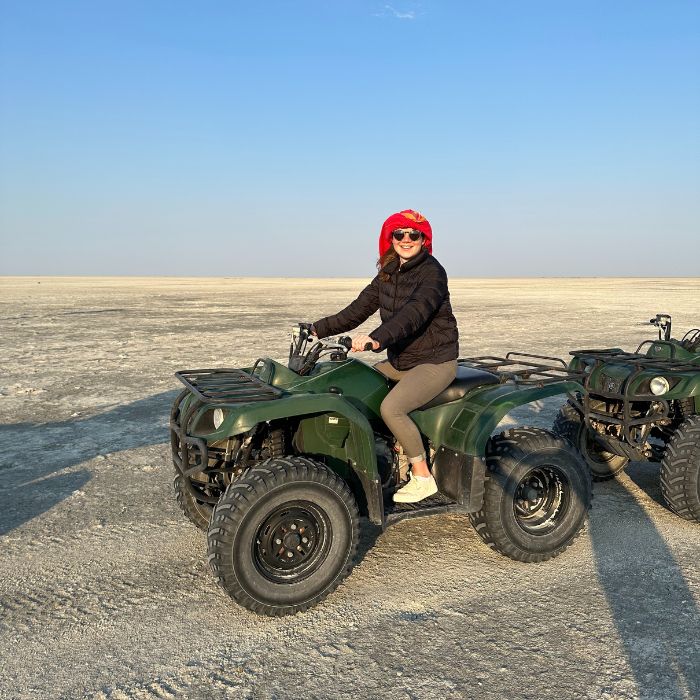 A woman wearing a red hat and sunglasses sits on a green ATV in a vast, barren landscape under a clear blue sky.