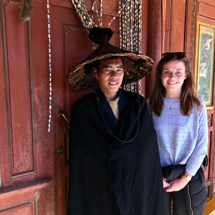A woman wearing a traditional hat and cloak stands next to a smiling girl in a casual sweater, both positioned in front of a colorful wooden door adorned with decorative beads.
