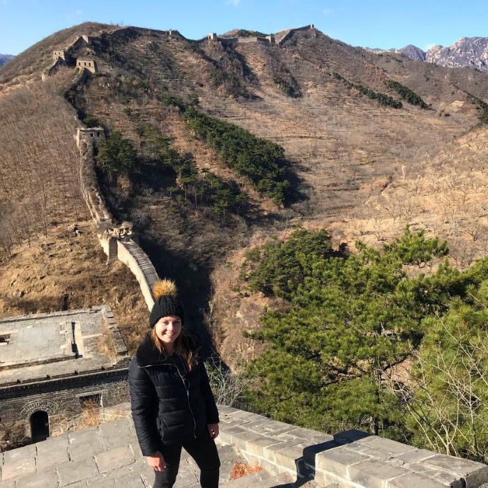 A person stands on a stone wall of the Great Wall of China, surrounded by mountainous terrain. The wall stretches into the distance, winding over hills in a mostly barren landscape with sparse trees.
