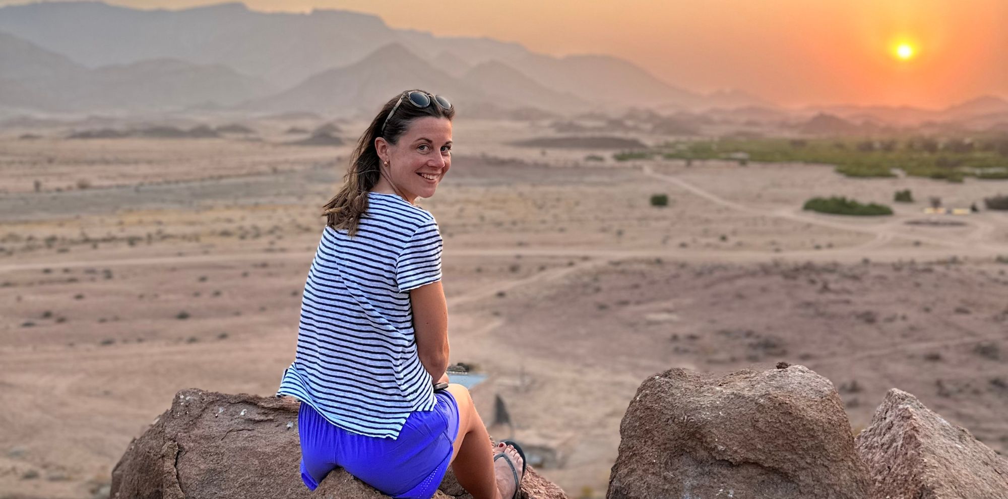 A woman in a striped shirt and blue shorts sits on a rock, smiling back at the camera. She is positioned against a backdrop of a sunset over a vast arid landscape with mountains in the distance.