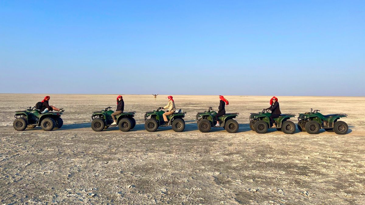 A line of six people on green ATVs, each wearing red headscarves, riding across a vast, dry, flat landscape under a clear blue sky.