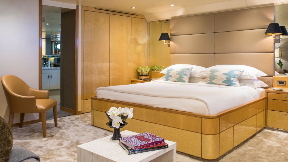 A stylish yacht cabin featuring a double bed with decorative blue cushions, wooden cabinetry, a plush carpet, and a modern armchair. Soft lighting creates a warm ambiance, complemented by a small table with decorative books and a plant.