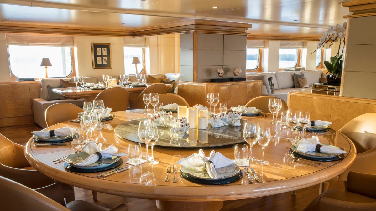 An elegant dining area featuring a large round table set for a meal. The table is adorned with fine glassware and neatly arranged cutlery, surrounded by stylish chairs. Soft lighting creates a warm atmosphere, complemented by decorative elements and a view through the windows.