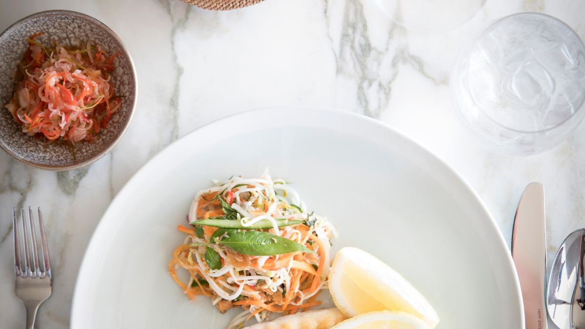 A beautifully arranged plate features grilled fish alongside a fresh salad made with carrots, cabbage, and snap peas, garnished with lemon wedges. A small bowl of pickled vegetables accompanies the dish, and elegant glassware is present on a marble table.