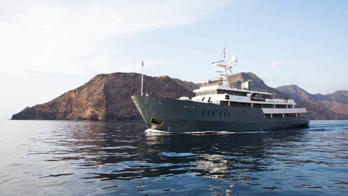 A luxury yacht cruising calmly on clear waters near a rugged, mountainous coastline.