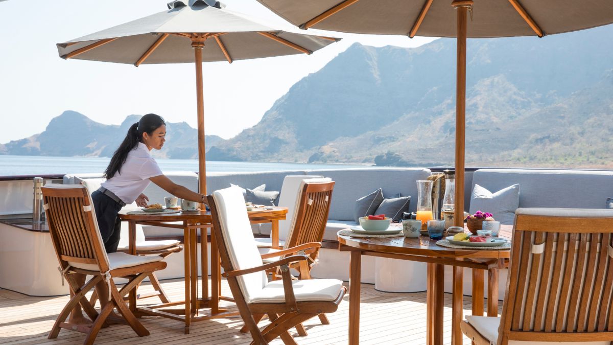 A woman sets a breakfast table on a sunny terrace overlooking a scenic mountain view. The table is adorned with plates, cups, and a vase of flowers, while shaded by a large umbrella.