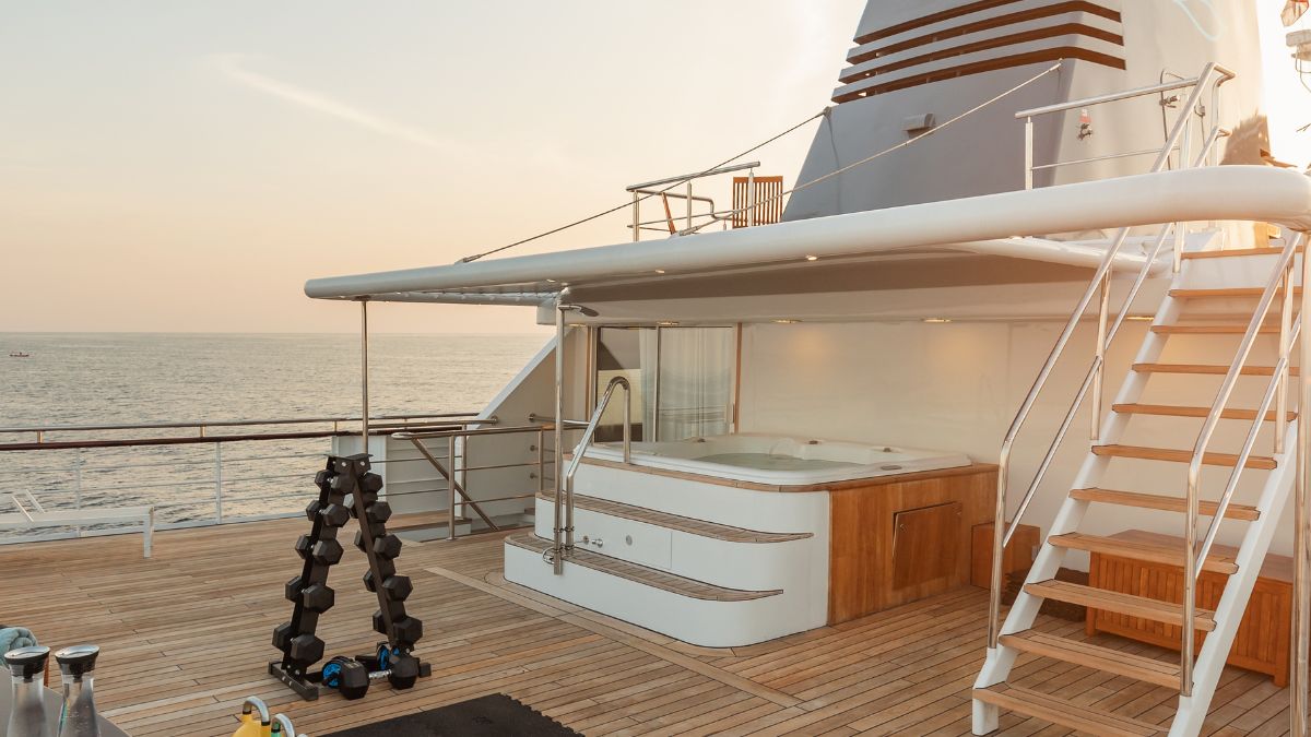 A luxurious yacht deck featuring a hot tub, outdoor furniture, a set of dumbbells, and a wooden staircase leading to additional levels, with a serene ocean view in the background.