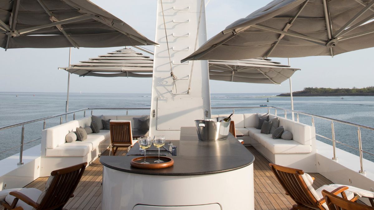 A luxurious yacht deck featuring comfortable seating with cushions, a central bar area, and an ice bucket. Large umbrellas provide shade while the sea glistens in the background.