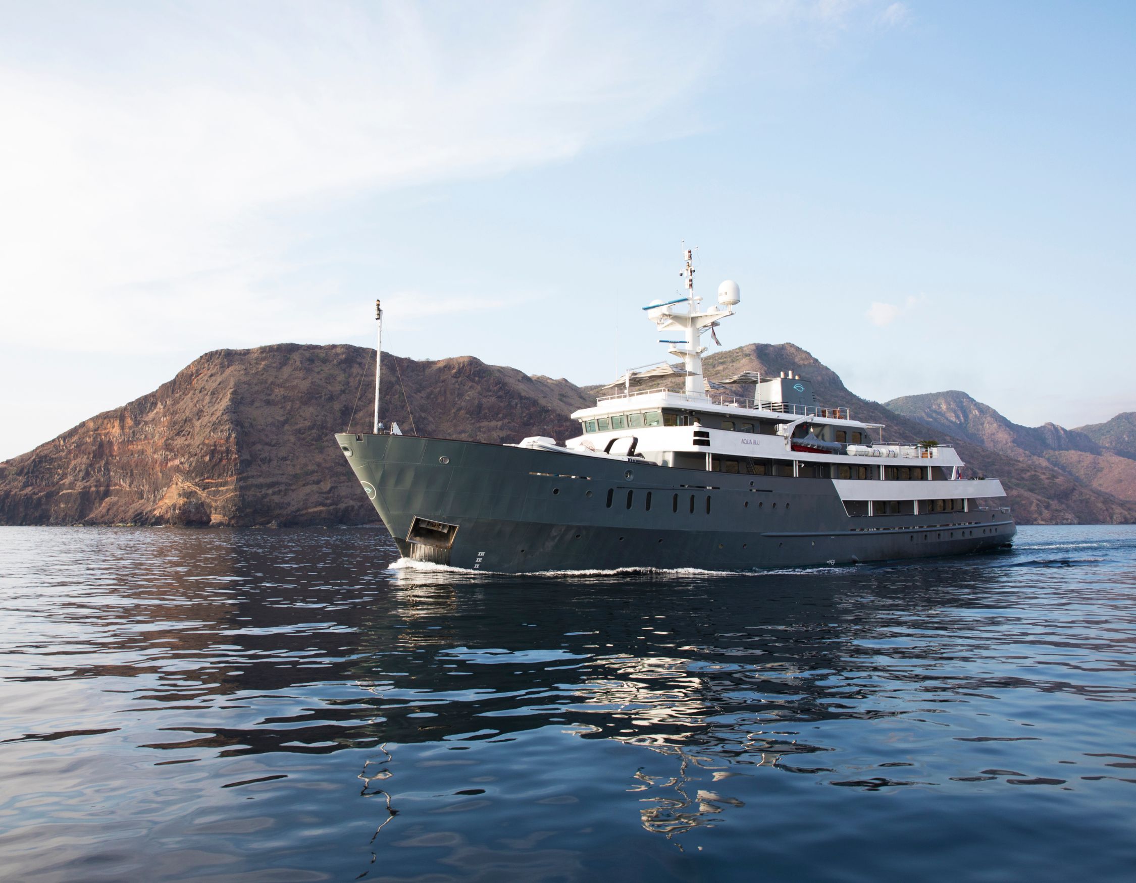 A sleek, modern yacht cruising on calm waters with rocky hills in the background. The yacht features a dark green hull and multiple decks, reflecting the afternoon sunlight.