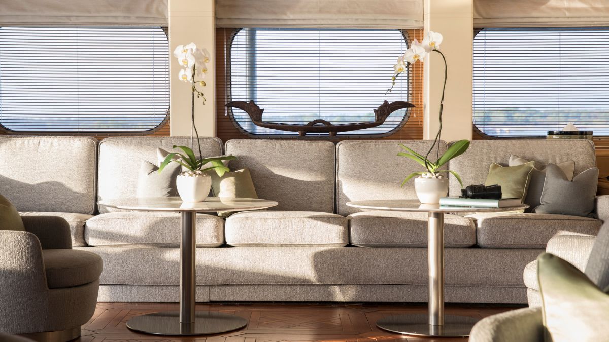 A cozy yacht interior featuring a light gray sofa with decorative pillows, two round side tables with plants in white pots, and a wooden sculpture in the background, illuminated by natural light from large windows.