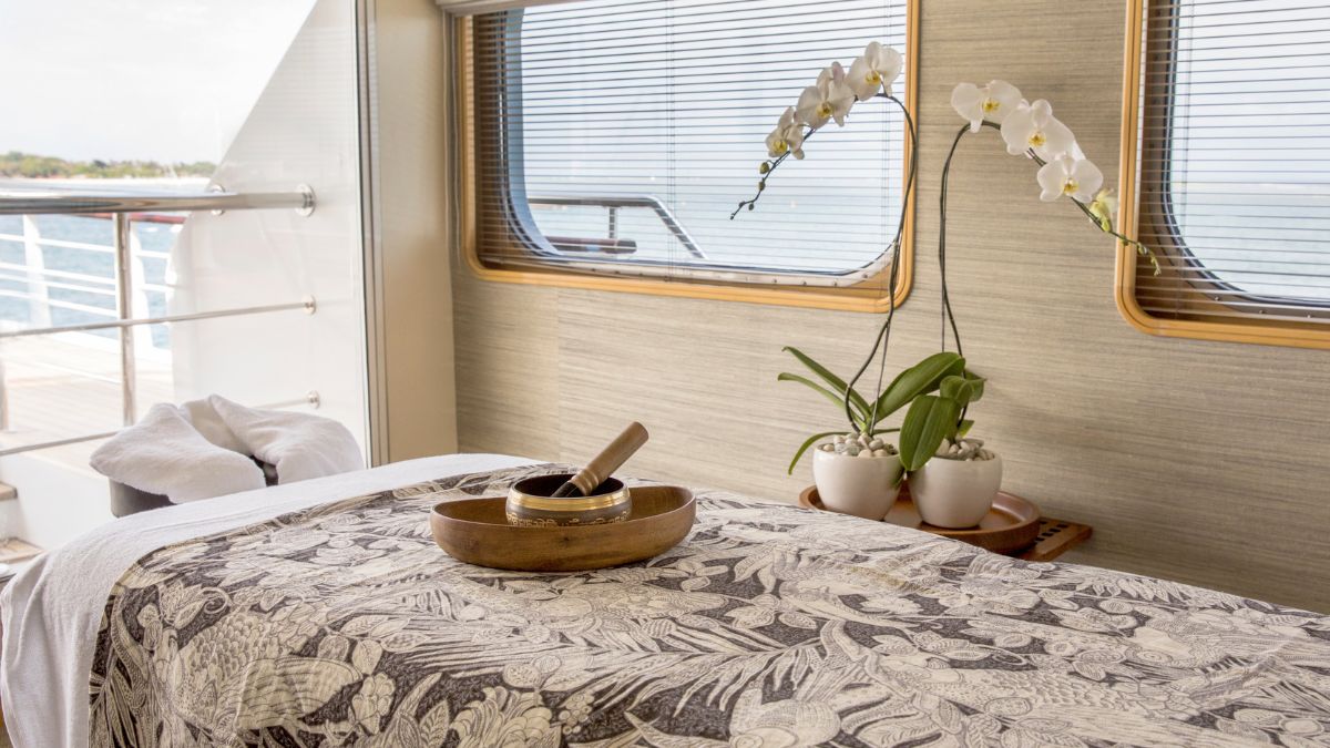 A serene spa setting featuring a massage table adorned with a patterned cloth. Nearby, two potted orchids add a touch of elegance, while a wooden bowl and pestle sit ready for use. Natural light filters through large windows, offering a view of the sea.