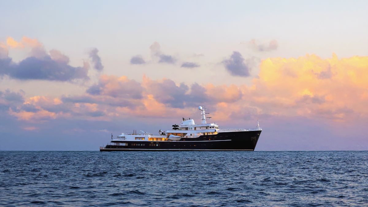 Large dark-hulled motor yacht anchored on a calm sea under a purple and orange sunset sky.