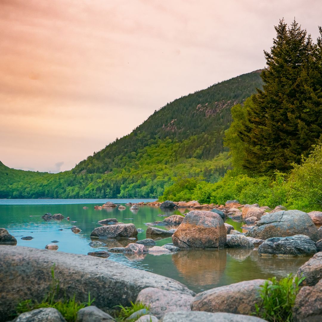 A tranquil lake surrounded by green hills and trees, with smooth boulders lining the shore. The sky displays soft pastel colors as the sun sets.