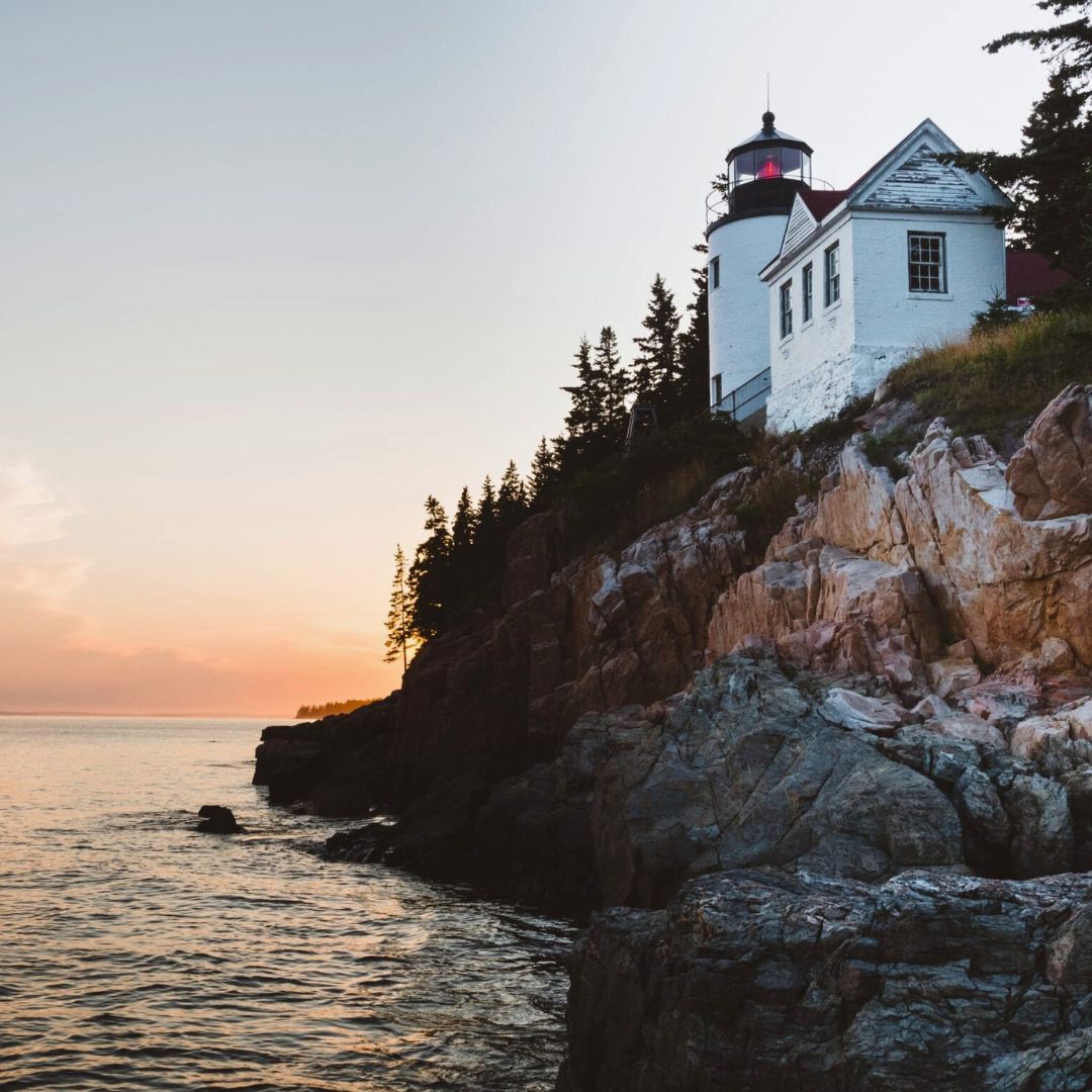 A lighthouse perched on a rocky coastline at sunset, surrounded by tall trees. The sky is painted in soft hues of orange and pink, reflecting on the calm ocean waters.