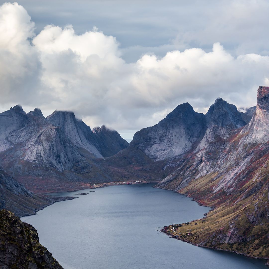 A serene fjord surrounded by rugged mountains under a cloudy sky. The water reflects the landscape, with a small village visible along the shore.