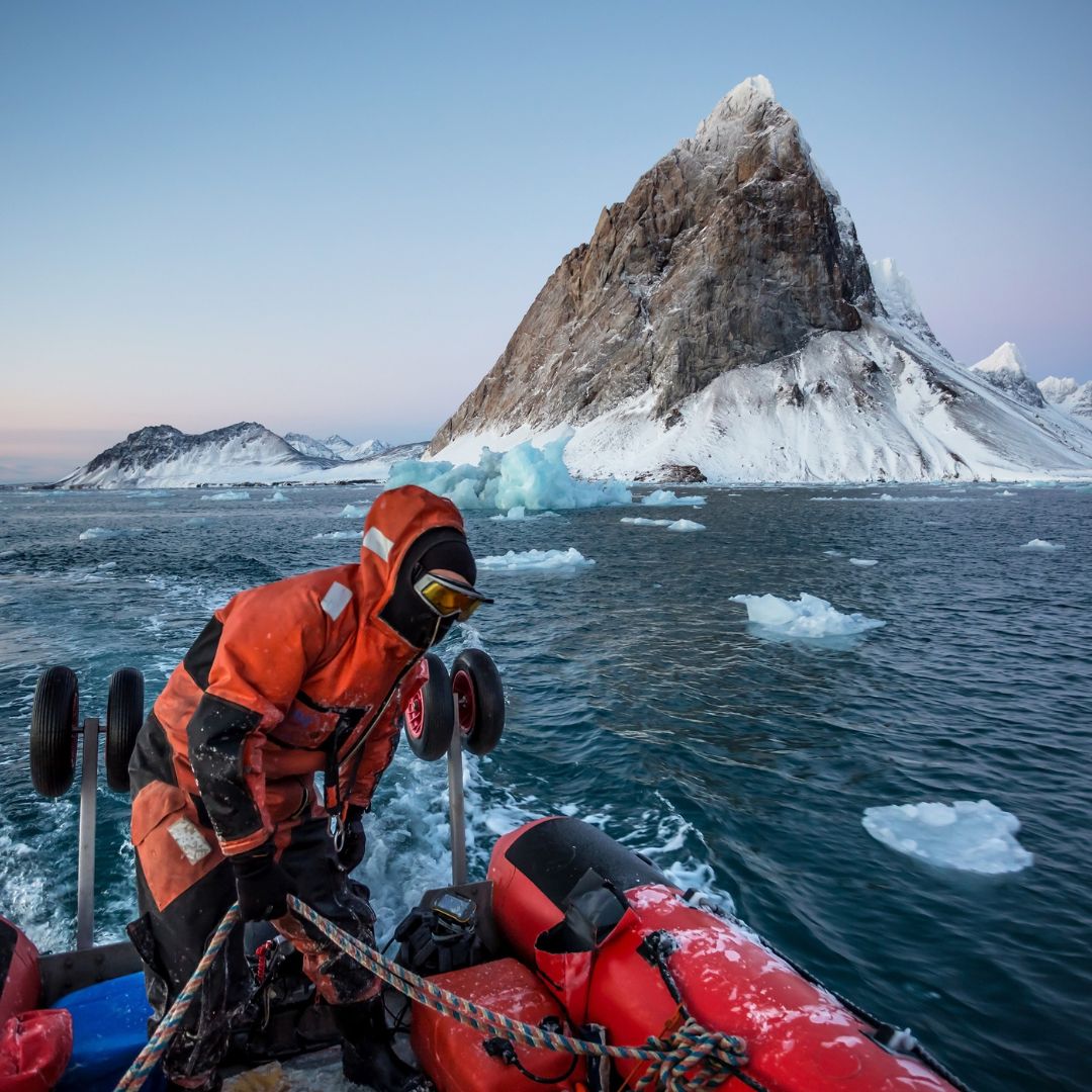 A person in an orange cold-weather suit prepares a small boat near an icy shore, with a large, snow-covered mountain looming in the background. The water is dotted with icebergs under a clear sky.