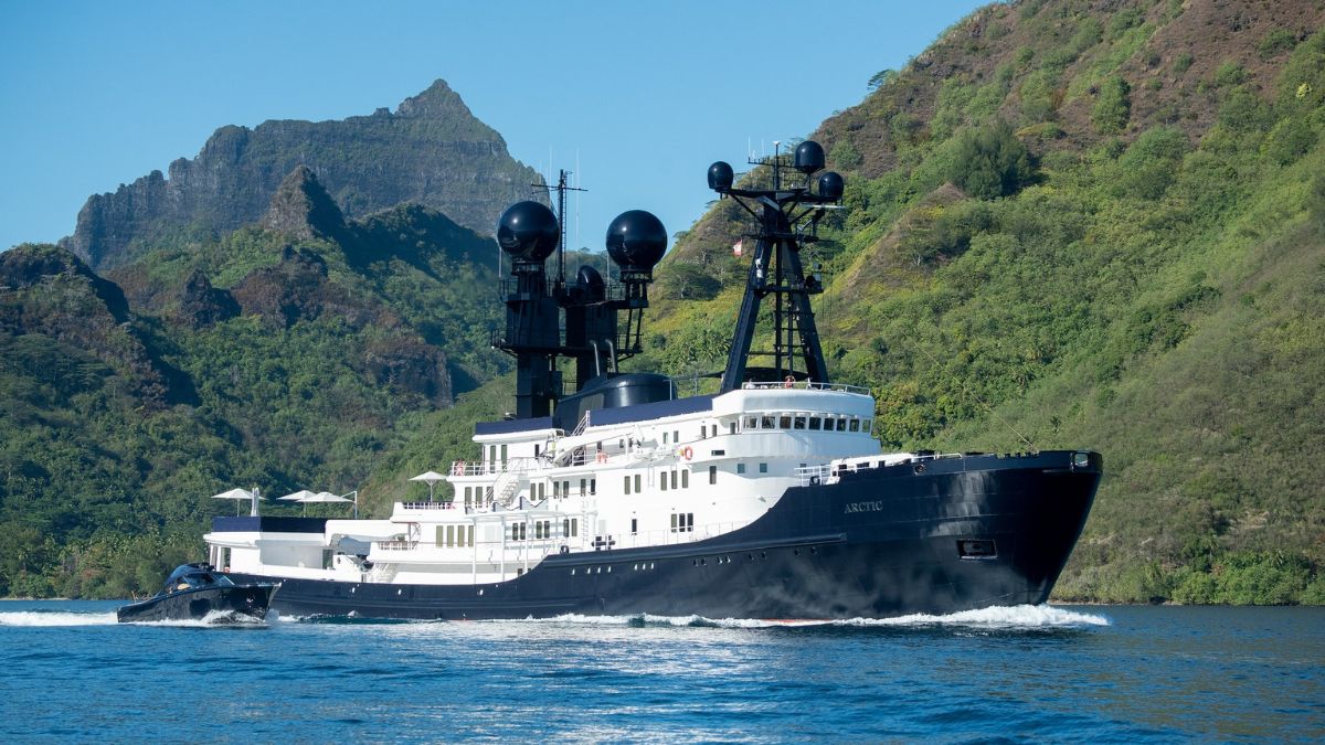 Large exploration yacht with black and white hull cruising past lush, steep green mountains..