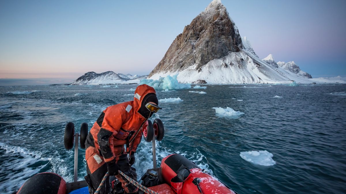 A person in an orange waterproof suit operates a small boat in icy waters, with a snow-covered mountain and icebergs in the background. The sky is tinted with soft pastel colours as the sun sets.