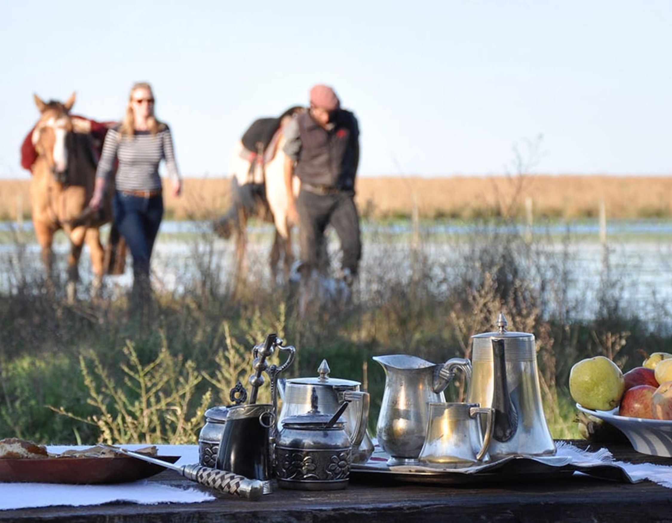 A rustic table is set with silver teapots and a platter of apples, while two people walk behind with horses near a tranquil body of water and grassy landscape.
