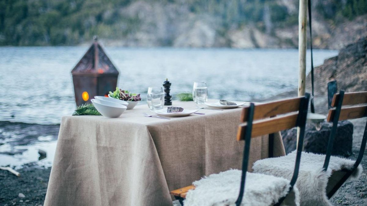 A beautifully set outdoor dining table by a serene lake. The table is covered with a beige cloth and features glasses, a bowl of salad, and a decorative lantern nearby. Two wooden chairs with fur accents are arranged around the table. The calm water and lush greenery create a peaceful backdrop.
