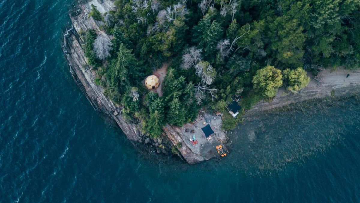 An aerial view of a rugged coastline with dense greenery, featuring a small clearing near the water's edge. There are tents set up on the shore, and a few kayaks are moored nearby. The area is surrounded by a mix of trees and rocky cliffs.