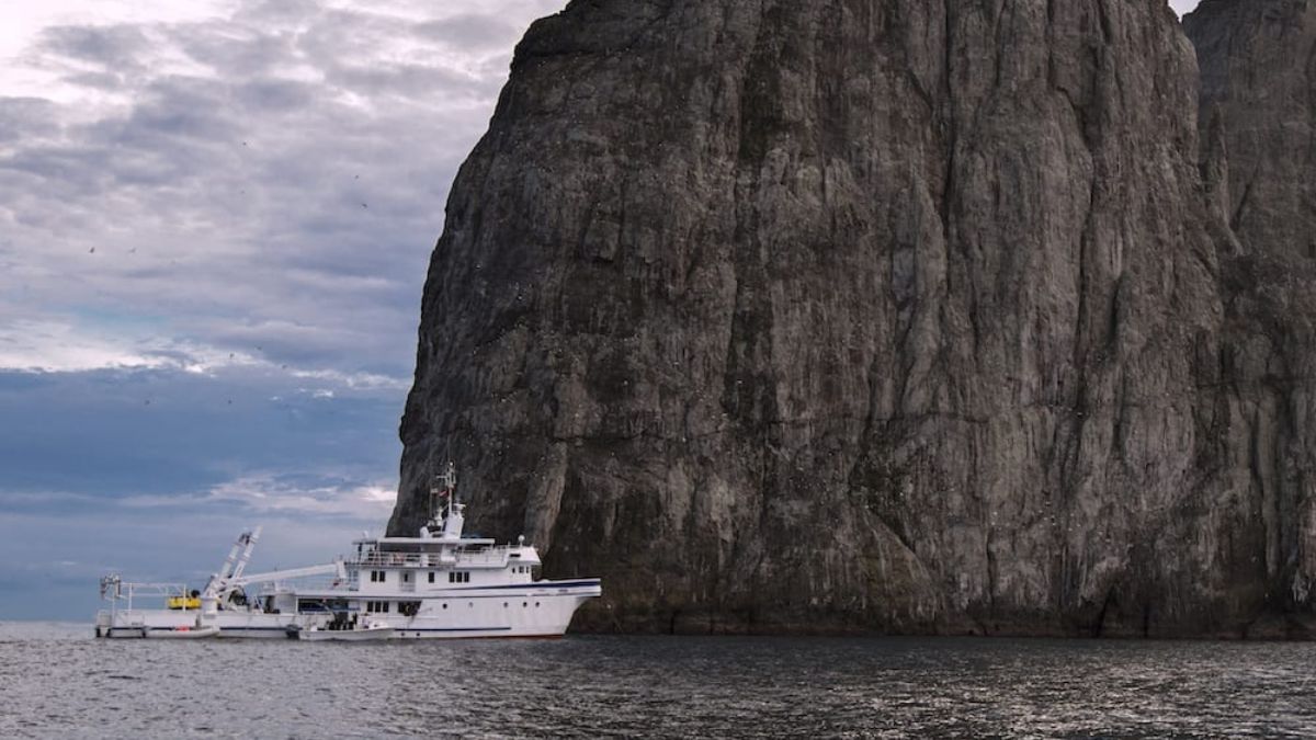 A large white research vessel anchored near a steep, rocky cliff under a cloudy sky. The boat is equipped with scientific gear and appears to be in a remote, natural setting.