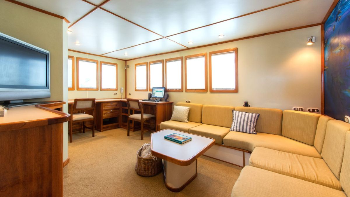 A comfortable lounge area featuring a curved yellow sofa, a small coffee table, and wooden accents. There is a television mounted on one wall and a desk with a computer in the corner, surrounded by large windows allowing natural light.