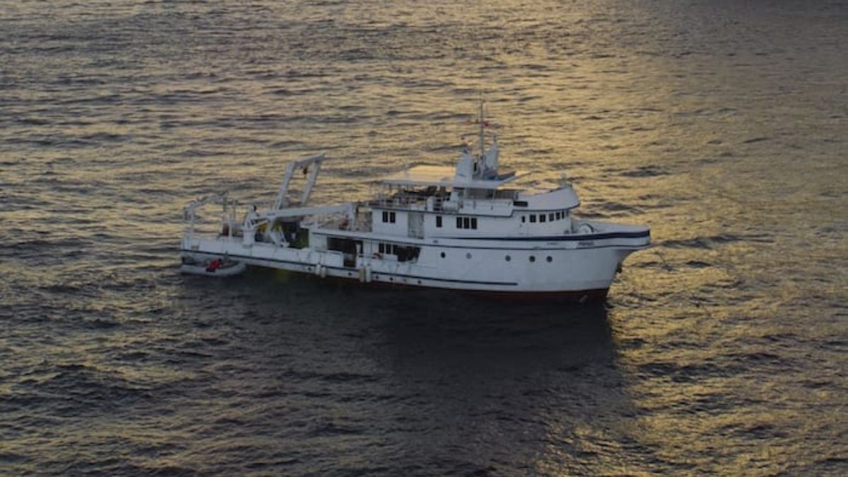 A large white ship with multiple decks floats on calm waters during sunset, reflecting golden hues.