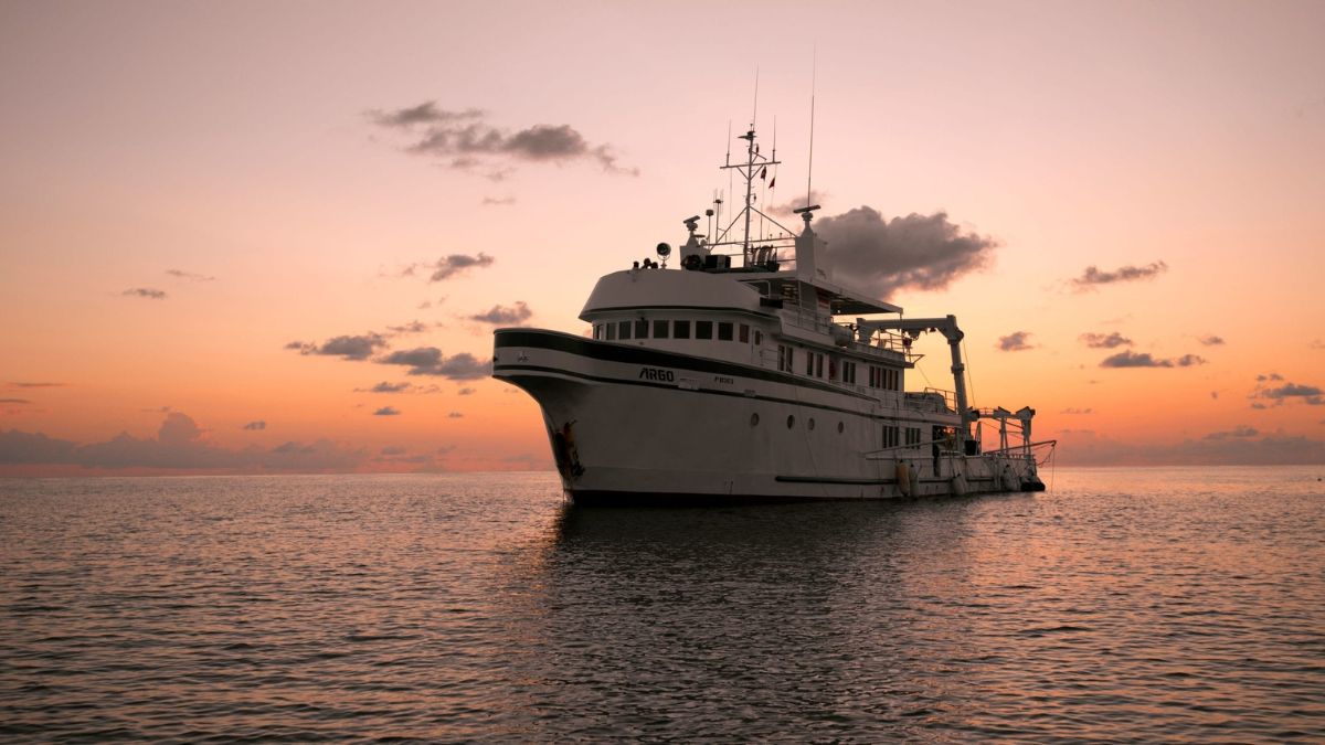 A large fishing vessel is anchored on calm water at sunset, with a vibrant orange and pink sky reflecting in the surface.