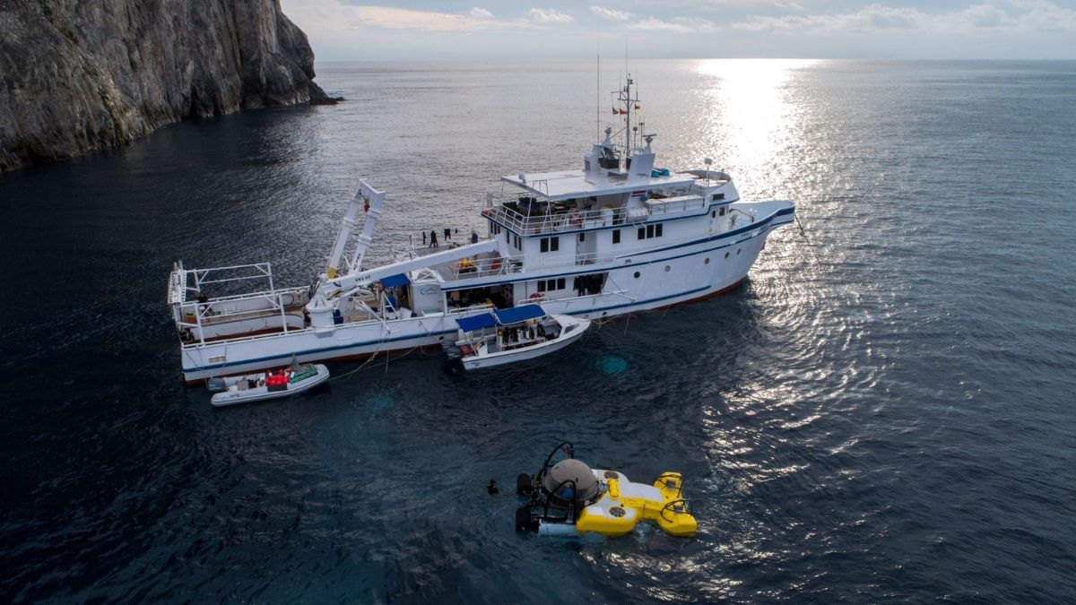 A large research vessel floats on calm waters near a rocky coastline, with a yellow submersible positioned beneath it. The sun reflects off the water's surface, creating a serene atmosphere.