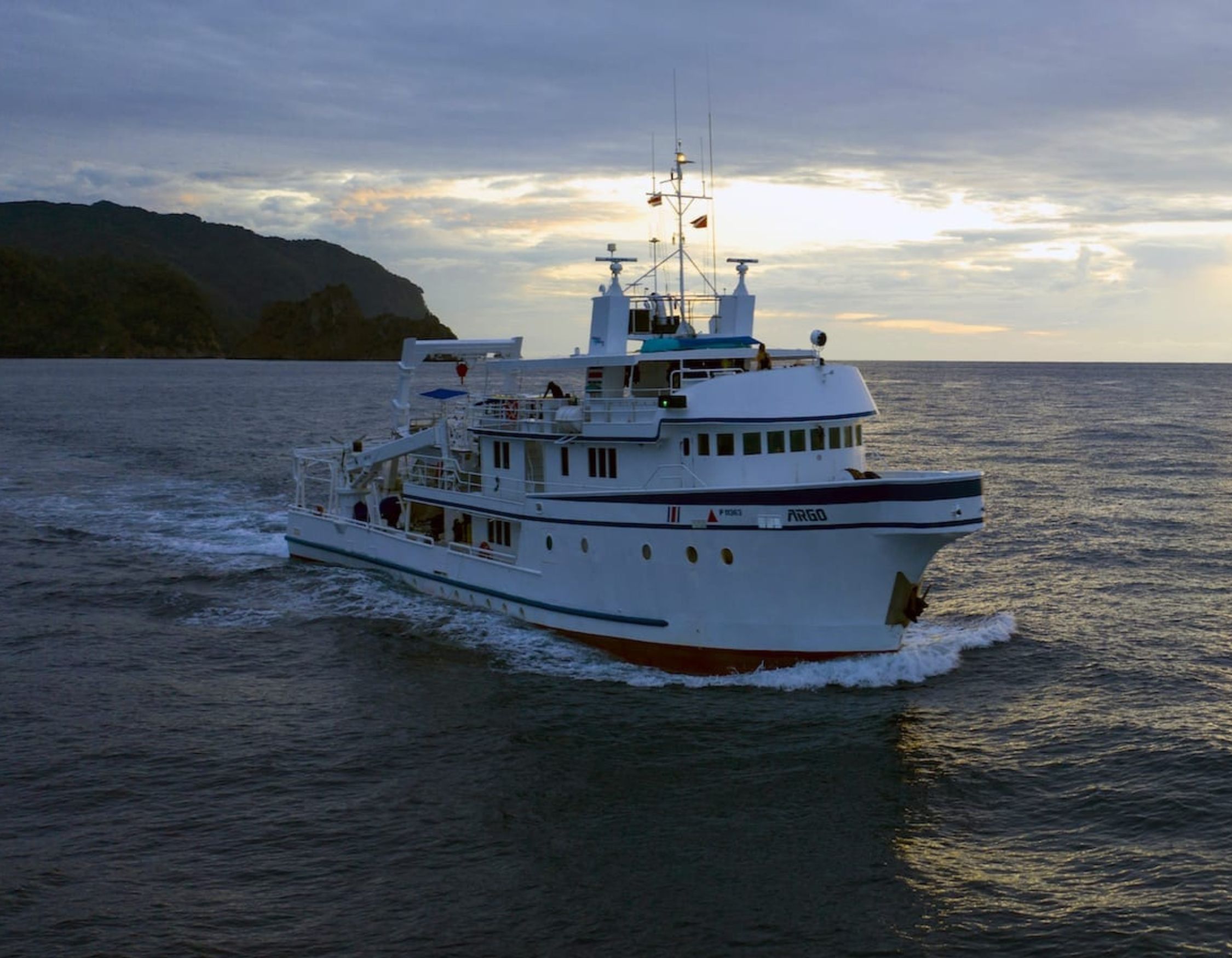 A large white and blue boat navigates through calm waters, leaving a trail of wake behind. The sky is partly cloudy with the sun setting in the background, illuminating the scene with golden light.