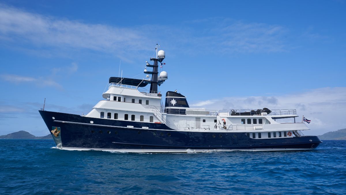 A large luxurious yacht with a blue and white hull sailing on clear blue water, surrounded by distant mountains and a blue sky.