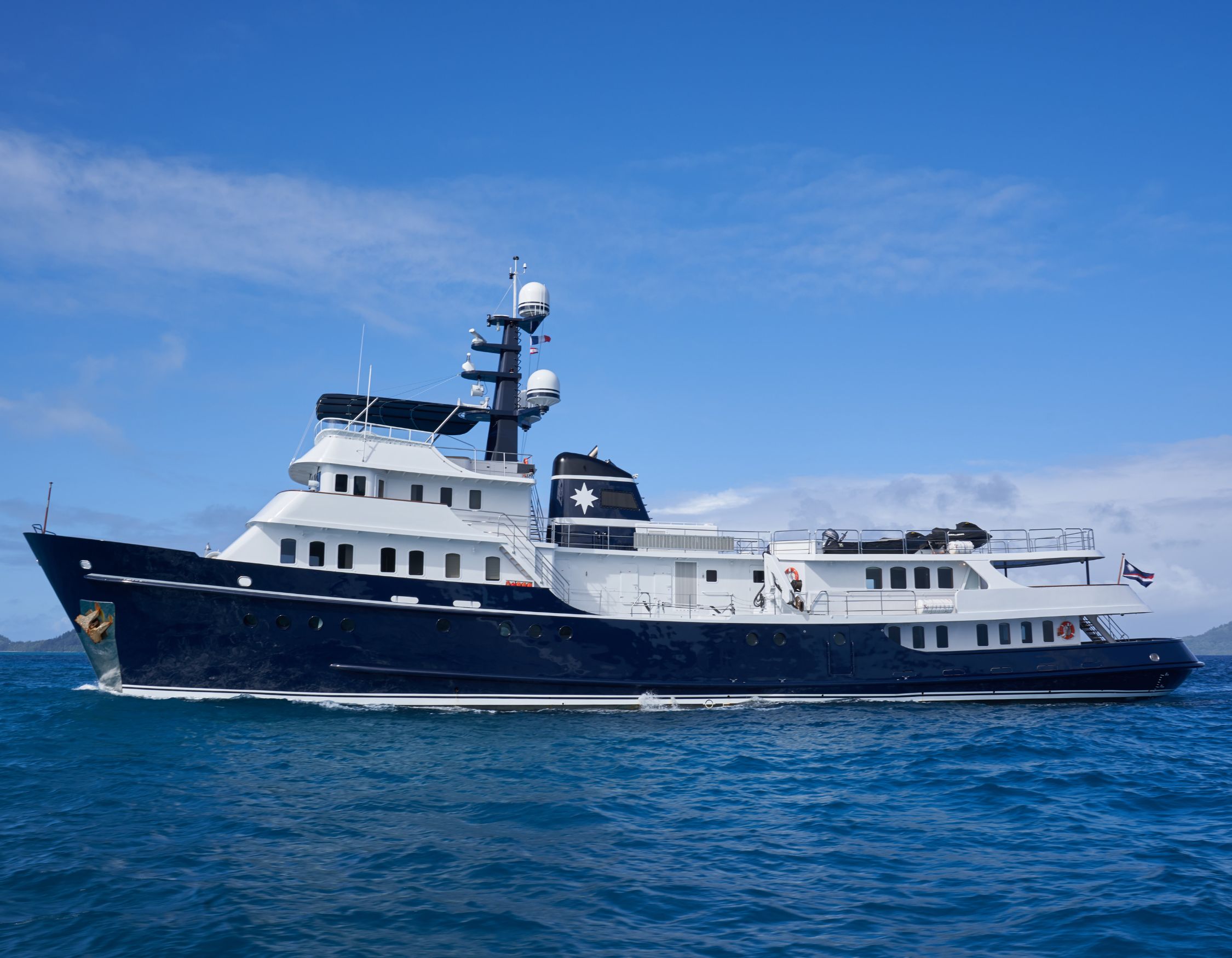 A large luxury yacht with a dark blue hull and white superstructure, featuring multiple decks and antennas, gliding through turquoise water under a clear blue sky.