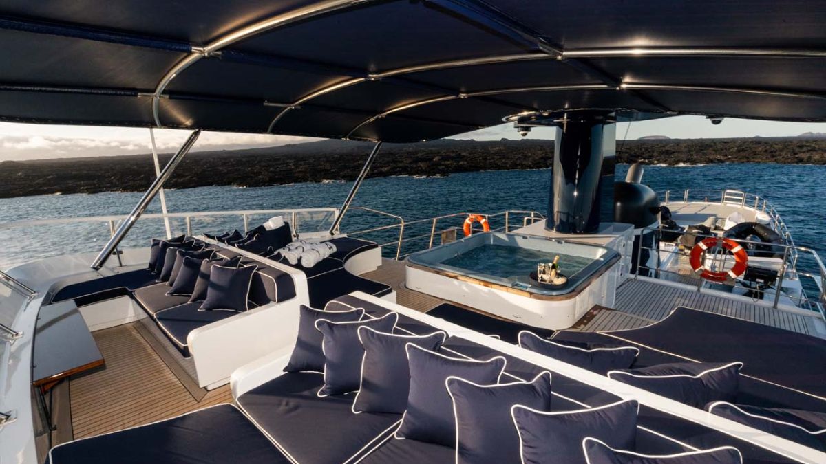 Luxurious yacht deck featuring spacious seating with navy cushions and a hot tub, overlooking calm waters. A scenic coastline is visible in the background under a clear sky.