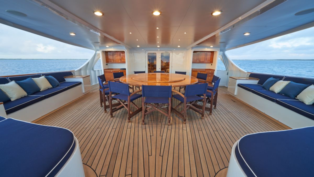 A spacious yacht interior featuring a circular wooden dining table surrounded by chairs, with blue cushions on the seating areas along the walls. Large windows offer views of the sea, and wooden flooring adds a touch of elegance.