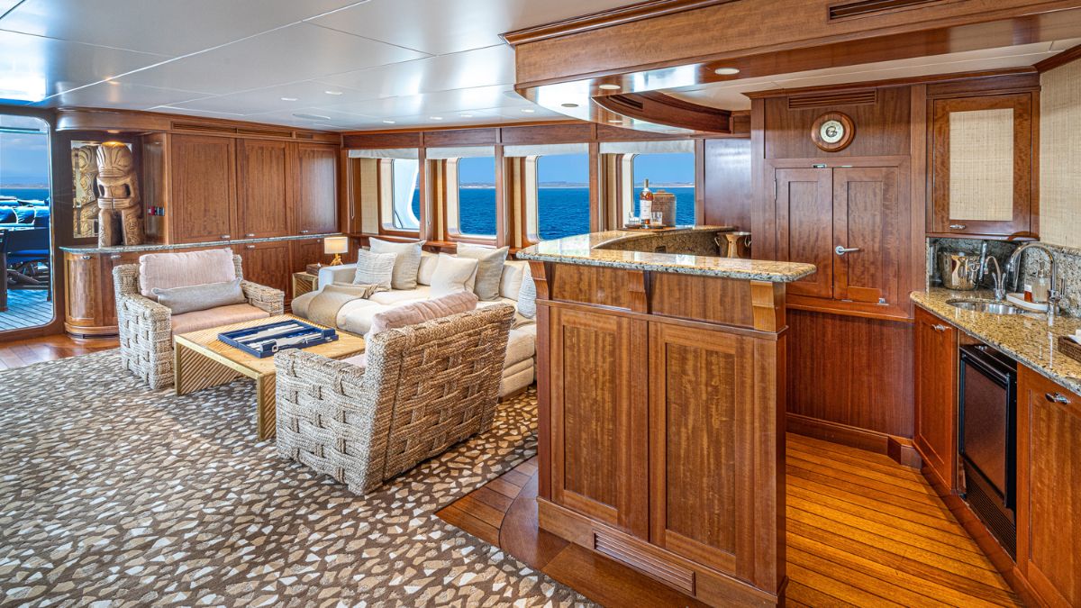 A luxurious interior of a yacht featuring a cozy living area with plush sofas, a wooden coffee table, and an open kitchen with granite countertops. Large windows provide an ocean view, and the warm wood paneling adds elegance to the space.