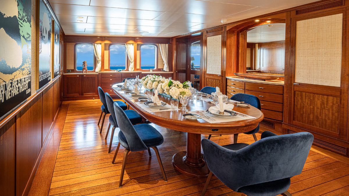 Elegant dining room on a yacht featuring a large wooden table set for a formal meal, surrounded by plush dark blue chairs. Fresh white flowers decorate the centre, and windows provide a panoramic view of the ocean. Warm wooden tones create a luxurious ambiance.
