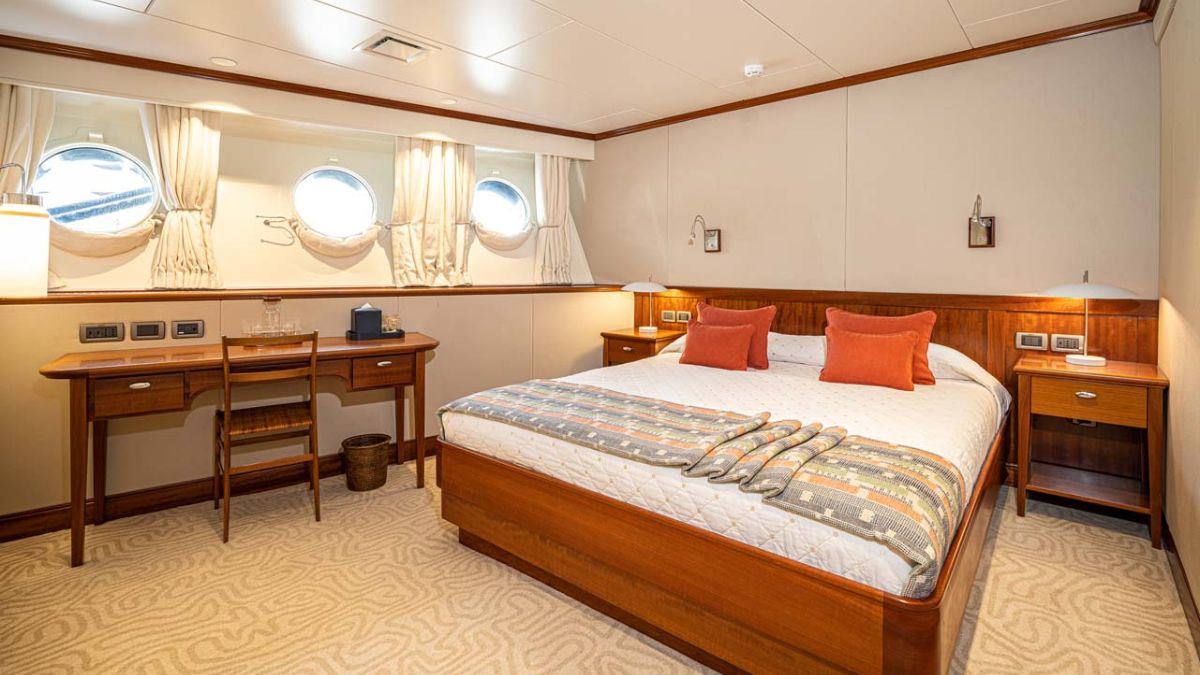A cozy bedroom featuring a queen-sized bed with orange accent pillows, a wooden desk and chair, and round porthole windows letting in natural light. The walls are neutral, with elegant decor and a plush carpet.