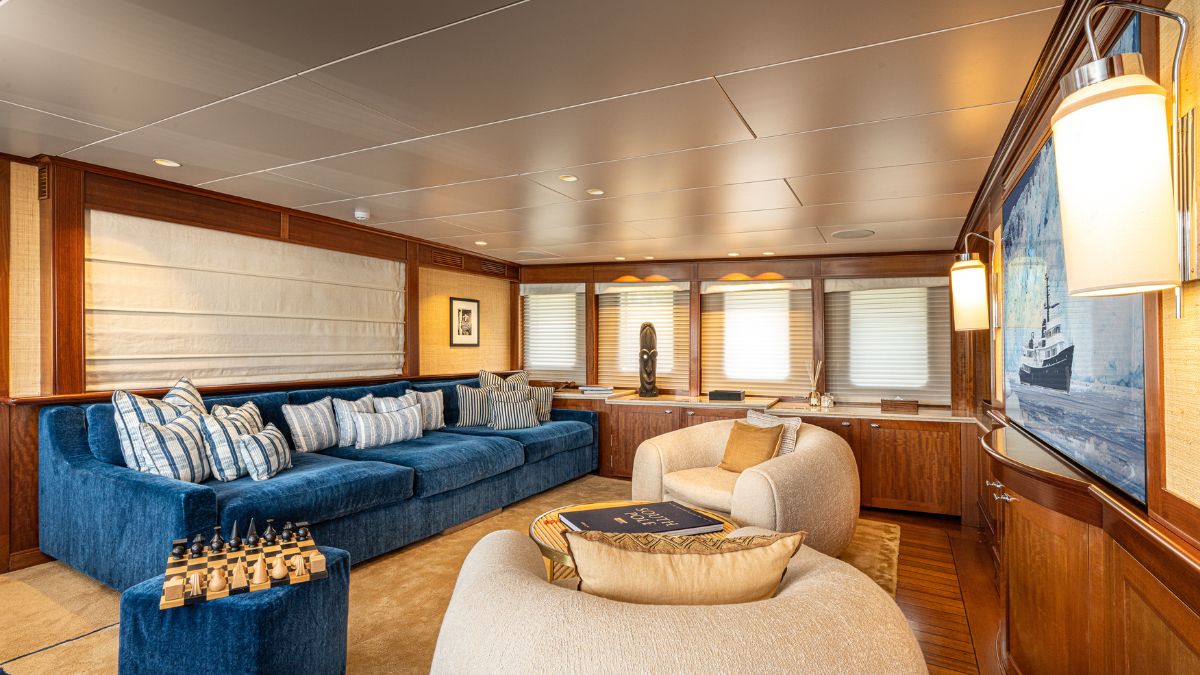 A luxurious yacht interior featuring a spacious living area with a blue sectional sofa adorned with decorative pillows, a light-colored curved chair, and a wooden desk with a view through large windows. A modern art piece and decorative items enhance the elegant ambiance.