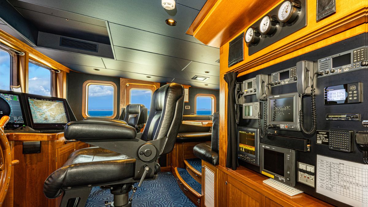 A captain's cabin on a ship featuring a large leather ergonomic chair, multiple screens displaying maps and navigational data, and various controls mounted on a wooden console. Large windows provide a view of the ocean.