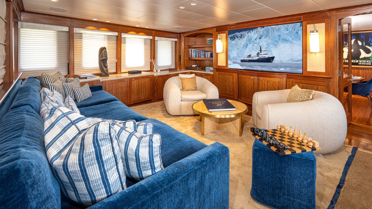 A spacious and elegantly designed living area featuring a large blue sectional sofa with decorative pillows, a round wooden coffee table, and a unique chess set. Soft lighting illuminates the room, which includes wooden accents and artwork on the walls, creating a cozy yet sophisticated atmosphere.
