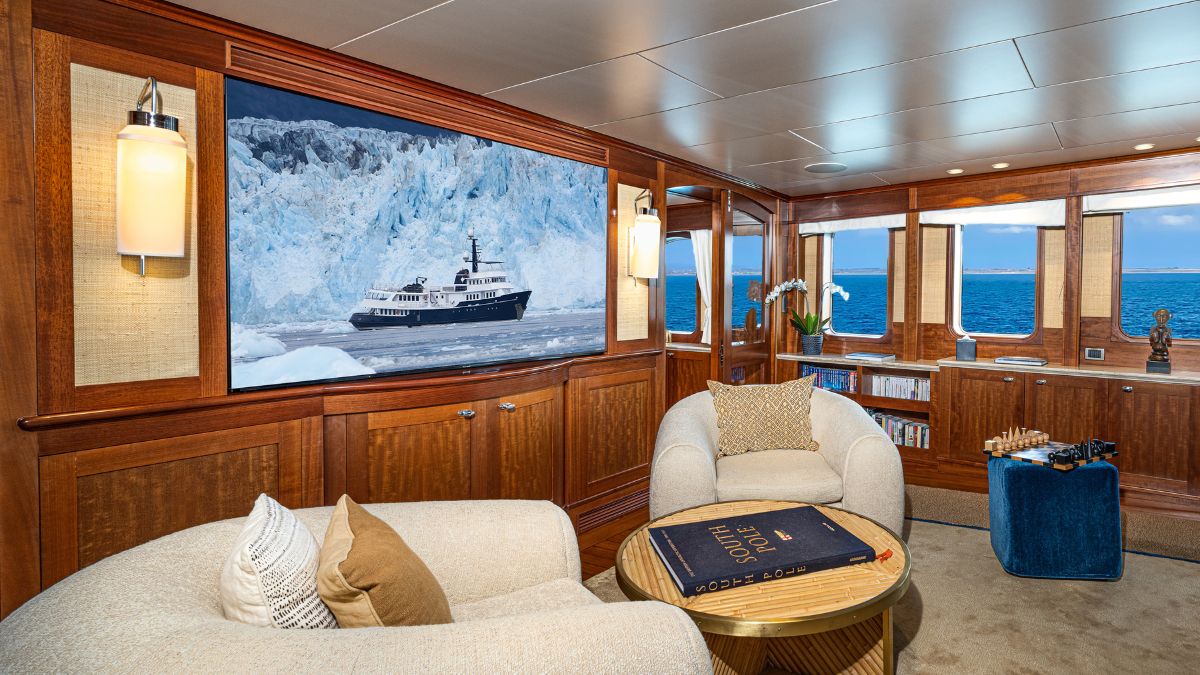A luxurious yacht interior features plush seating, wooden paneling, and a large screen displaying a ship near a glacier. A coffee table with an open book and a small blue stool adds to the elegant decor. Ocean views are visible through large windows.