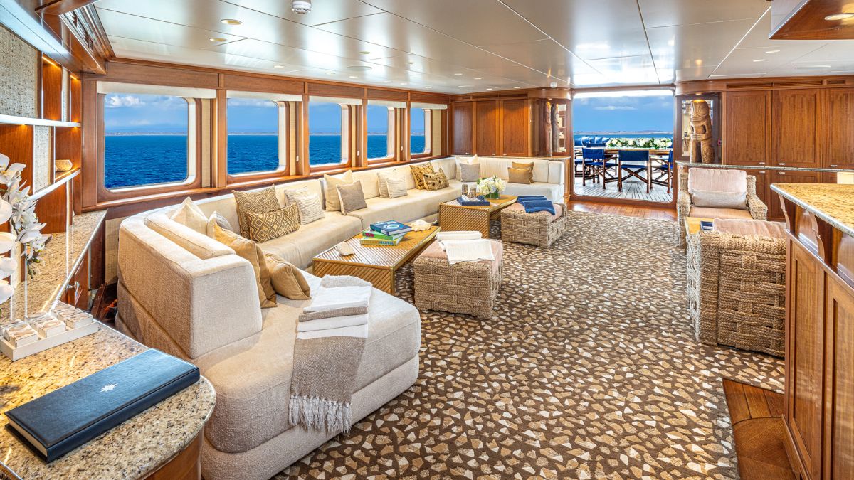 A spacious and elegantly decorated yacht interior featuring a large, cozy sectional sofa, woven armchairs, and natural light streaming through wide windows. The room has a light color palette, with neutral tones and textured fabrics, and a view of the ocean visible through the glass.