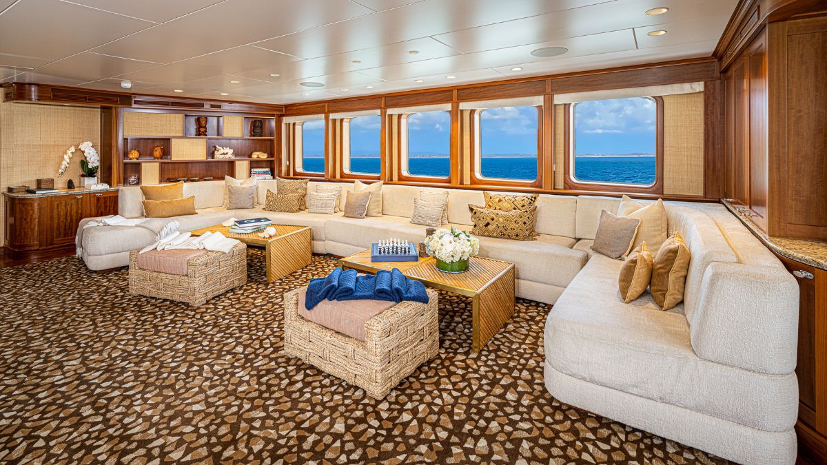 Luxurious boat interior featuring a spacious lounge with a large beige sectional sofa, decorative cushions, coffee tables, and floral arrangements. Large windows offer a view of the ocean.