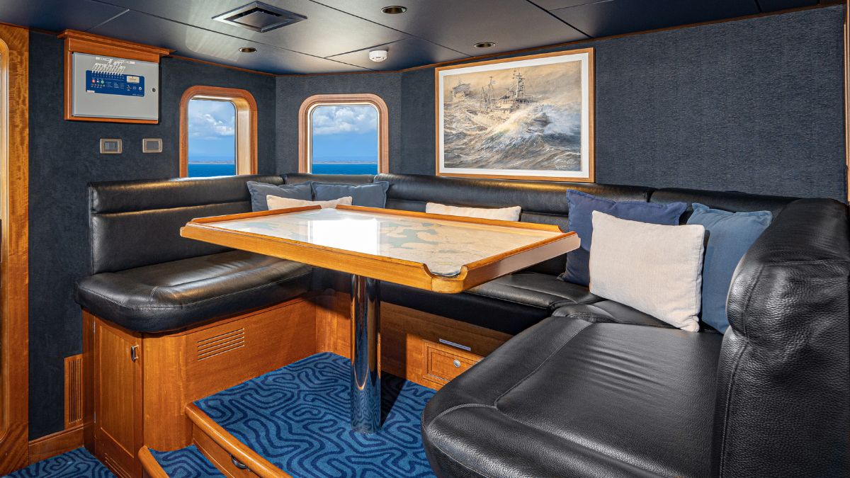 A cozy seating area in a boat, featuring a wooden table with a map on it, surrounded by dark leather couches and decorative pillows. Large windows provide a view of the ocean and a framed artwork of turbulent seas hangs on the wall.